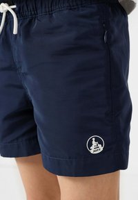 Pantaloni da bagno blu navy realizzati in tessuto leggero, con vita elasticizzata e cordoncino, e un dettaglio con logo bianco sull'orlo inferiore.