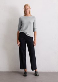Jasnoszary sweter z długim rękawem i dekoltem w łódkę, zestawiony z dopasowanymi, ciemnoniebieskimi spodniami o szerokich nogawkach. Model nosi czarne buty na slip-on.
