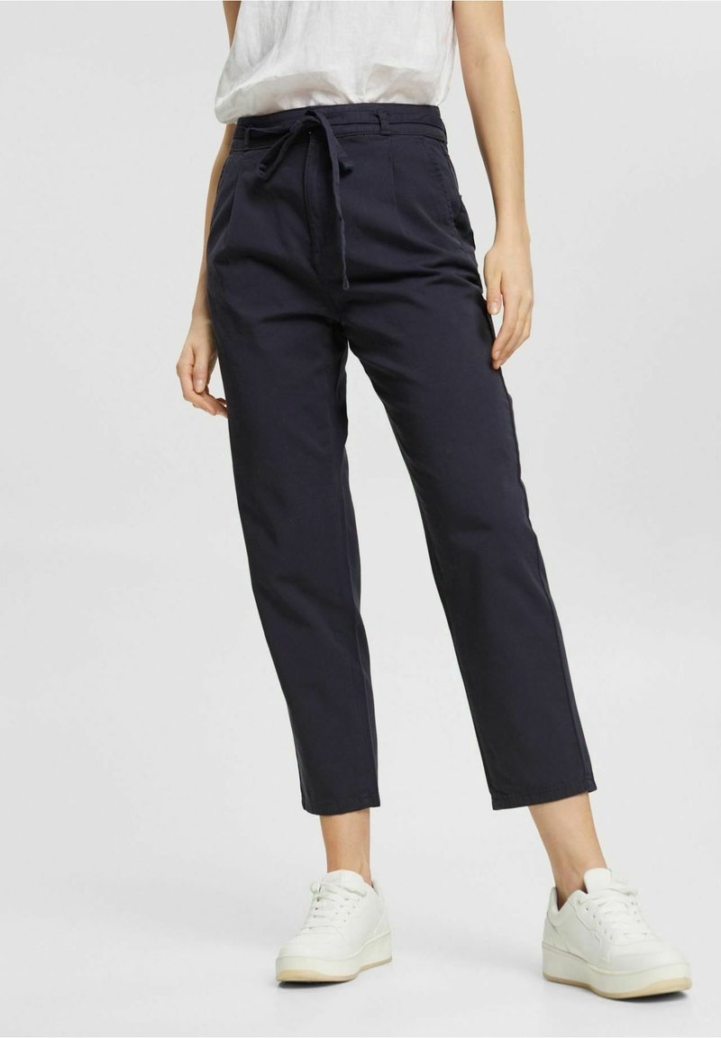 Personne portant un pantalon bleu marine à cordon, associé à des baskets blanches et un haut blanc ample, debout devant un fond clair uni.