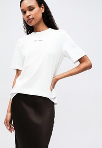 White cotton t-shirt with short sleeves, featuring a minimalist design and center logo, paired with a dark brown satin skirt.