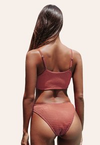 Costume da bagno a due pezzi color ruggine con texture a coste, caratterizzato da un top corto con spalline sottili e un bikini a vita alta. Texture liscia, design minimale.