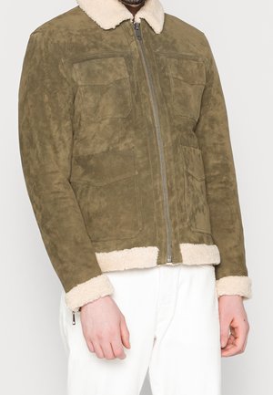 Mann trägt olivgrüne Wildlederjacke mit vier vorderen Klapptaschen, Lammfellkragen und -manschetten, weiße Hose, steht vor weißem Hintergrund.