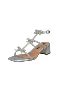 Sandalo con tacco quadrato in argento, dotato di tre fiocchi decorativi e una fascia adornata di strass. Presenta un tacco a blocco basso e una fibbia alla caviglia.