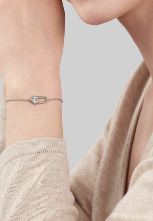 Zarter silberner Kettenarmband mit herzförmigem Schlossanhänger, getragen an einem erhobenen Handgelenk, beiger Strickärmel sichtbar.