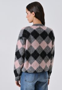 Donna di spalle che indossa un maglione peloso a losanghe nei colori rosa, grigio e nero, abbinato a jeans blu e orecchini dorati.