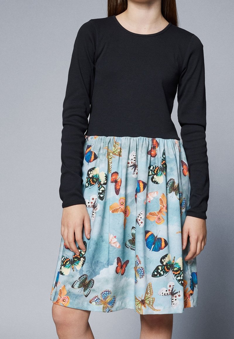 Fille portant une robe noire à manches longues avec une jupe bleue imprimée de différents papillons colorés.