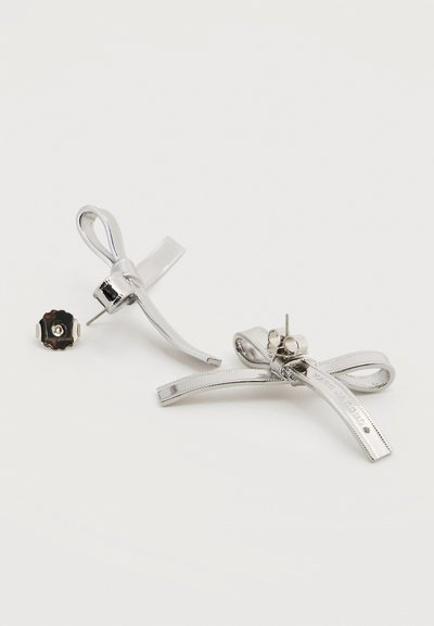 Ασημί μεταλλικά σκουλαρίκια καρφίτσες σε σχήμα φιόγκου με πεταλούδα στο πίσω μέρος, τοποθετημένα σε λευκή επιφάνεια.