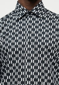 Κοντινό πλάνο ενός μαύρου πουκαμίσου με επαναλαμβανόμενο λευκό γεωμετρικό μοτίβο και μικρό κείμενο «Karl Lagerfeld» στο γιακά και στο μπροστινό μέρος.