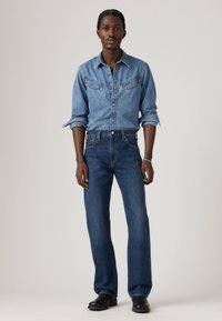 Μπλε πουκάμισο από τζιν με τσέπες στο στήθος και κούμπωμα με κουμπιά, συνδυασμένο με σκούρα μπλε τζιν καμπάνα και μαύρες δερμάτινες μπότες. Ουδέτερο φόντο.