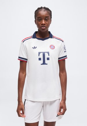 Personne aux cheveux tressés portant un maillot blanc du Bayern Munich et un short, debout sur un fond clair uni.