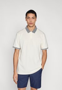 Κρεμ πόλο μπλούζα με γκρι γιακά και περιθώριο στα μανίκια, με κουμπωτό κλείσιμο και λογότυπο, συνδυασμένη με σκούρο μπλε σορτς. Λείο ύφασμα.