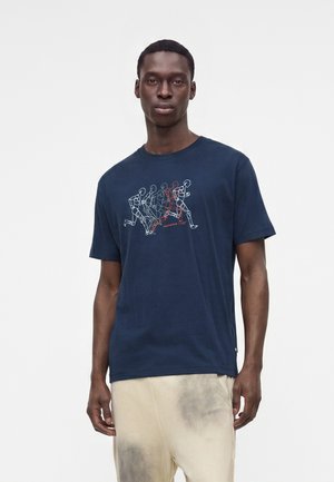 Homme portant un t-shirt bleu marine avec un motif graphique d'une séquence de personnages en course blancs et rouges et un pantalon beige clair avec des taches foncées.
