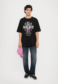 Black oversized t-shirt with colorful graphic design and text, paired with dark blue jeans and a pink cap featuring embroidered text.