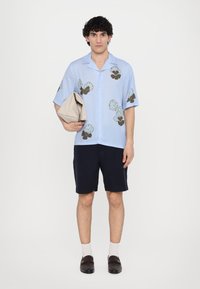 Lyseblå kortermet skjorte med grønne og brune blomster mønstre, parret med marineblå shorts. Modellen holder en beige veske og har på seg svarte slipsko.