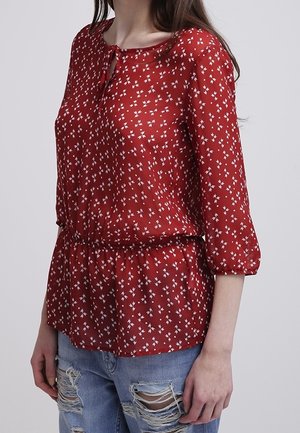 Frau trägt eine rote Bluse mit weißem Blumenmuster und Dreiviertelärmeln, kombiniert mit zerrissenen blauen Jeans, steht vor einem schlichten Hintergrund.
