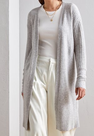 Cardigan lavorato a maglia grigio chiaro con maniche a coste, apertura frontale e una trama che combina motivi lisci e a coste. Abbinato a un top bianco aderente.
