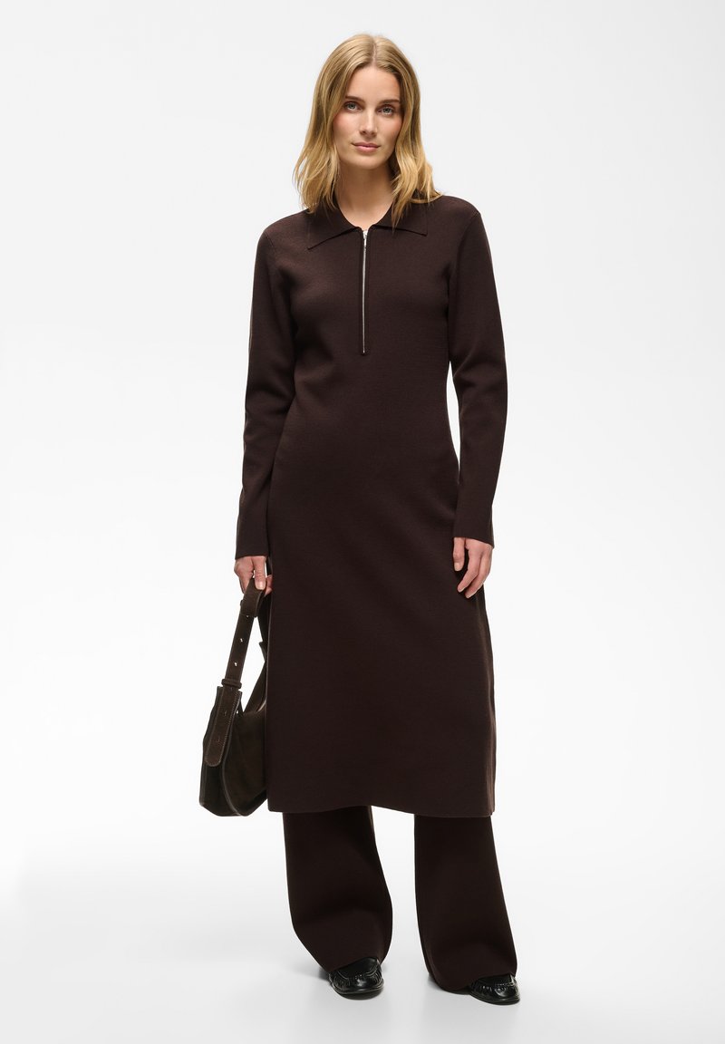 Femme blonde portant une longue robe zippée marron foncé par-dessus un pantalon large assorti, tenant un sac à main foncé, debout sur un fond blanc.