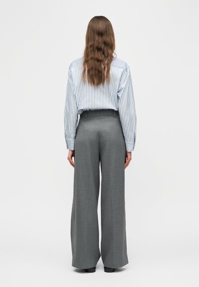 Femme aux longs cheveux bruns ondulés portant une chemise rayée bleu et blanc rentrée dans un pantalon gris taille haute à jambes larges, debout de dos.
