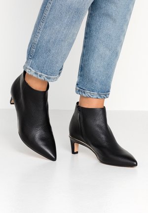 Pieds portant des bottines en cuir noir à bout pointu avec talons bas, associées à un jean denim bleu retroussé sur fond blanc.