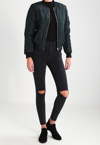 Mörkgrön bomberjacka med dragkedja fram, ribbade muddar och en dragkedjeficka på ärmen. Svarta tajta jeans med knäskär och svarta ankelboots.