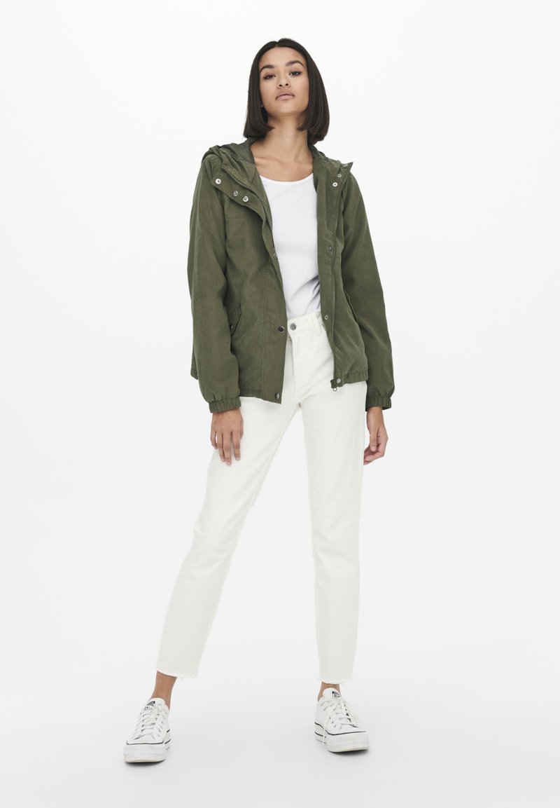 Olijfgroene hoodie met drukknoopsluitingen, gedragen over een witte top, gecombineerd met witte broeken en zwart-witte sneakers. Soepele textuur.