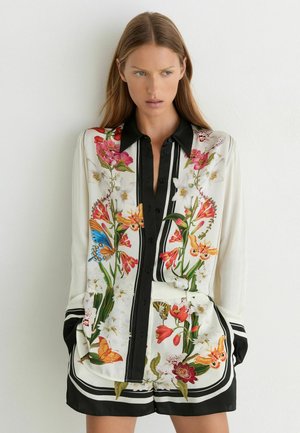 Vrouw met lang bruin haar die een wit shirt met bloemen- en vlinderprint draagt en bijpassende korte broek met zwarte rand, staand tegen een witte muur.