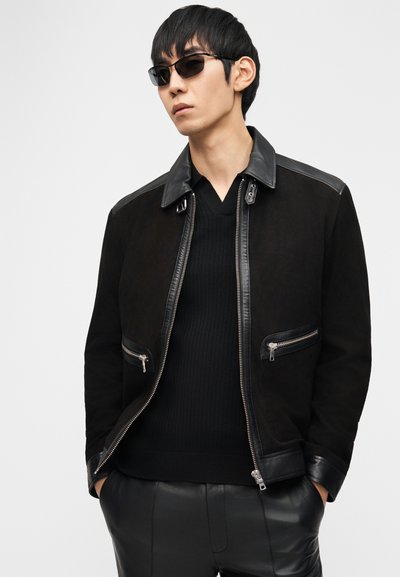 Άνδρας που φοράει μαύρο μπουφάν με λεπτομέρειες από δέρμα, μαύρο ριμπ πουλόβερ, μαύρο δερμάτινο παντελόνι και σκούρα γυαλιά ηλίου μπροστά σε λευκό φόντο.