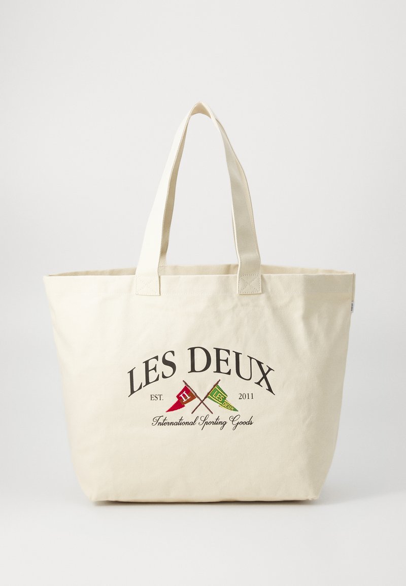 Canvas tote bag in cream with dual handles. Features embroidered text and graphic flags in red and green. Durable, spacious design.