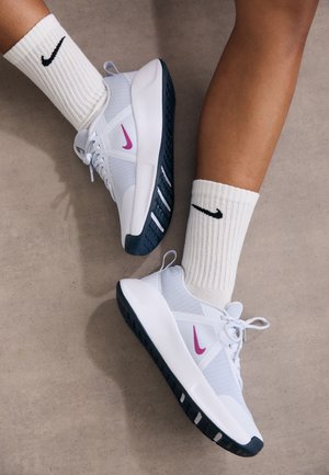 Beine mit weißen Nike-Socken und weißen Turnschuhen mit pinkfarbenen Nike-Logos und schwarzen Sohlen auf neutralem Boden.