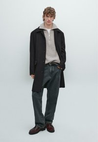 Svart kappa över en grå tröja med dragkedja i kragen, matchad med m�örka jeans och bruna skor. Modellen står med händerna i fickorna mot en ljus bakgrund.