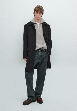 Cappotto nero su un maglione grigio con colletto a zip, abbinato a pantaloni jeans scuri e scarpe marroni. Il modello sta in piedi con le mani in tasca su uno sfondo chiaro.