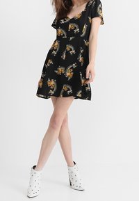 Svart kortärmad klänning med blommiga och paisleymönster, utsvängd fåll, längd ovanför knäna, ihop med vita ankelskor med metalliska nitar.