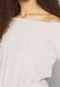 Pull en tricot beige clair à épaules dénudées avec une texture douce et une coupe décontractée, doté d'un large col et d'un motif côtelé subtil.