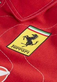 Logo giallo Ferrari con un cavallo nero in corsa e i colori della bandiera italiana su un tessuto rosso con cuciture bianche.