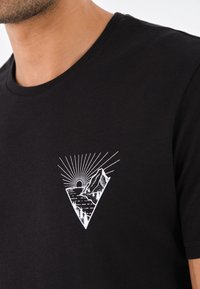 Czarny bawełniany t-shirt z białym trójkątnym motywem graficznym przedstawiającym góry i promienie słoneczne. Okrągły dekolt, krótkie rękawy, gładka tekstura.