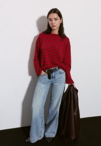 Red and black striped long-sleeve shirt paired with light blue wide-leg jeans. Carrying a brown leather jacket and wearing pointed, studded shoes.