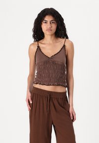 Brun cropped topp med justerbara spaghetti band, smockat tygdesign, volangkant längs halsringning och nederkant, bärs med matchande bruna byxor.