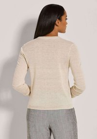 Frau mit langen dunklen Haaren, die ein beiges Langarmoberteil und graue Nadelstreifen-Hosen trägt, mit dem Rücken zu einer einfarbigen Hintergrund.