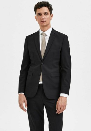 Svart dress med skreddersydd passform, enkel knappelukking, klaffelapper, to frontlommer, kombinert med et lyse farget slips og hvit skjorte.
