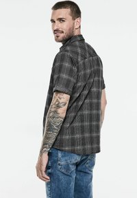 Košile krátkého rukávu černé a šedé kostkované s límečkem na knoflíky. Nosená s modrými džíny. Viditelné tetování na ruce.