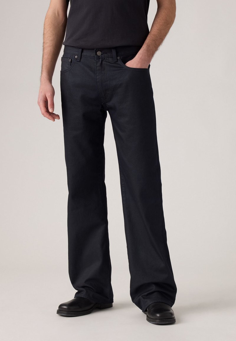 Man in losse zwarte jeans en zwarte schoenen, met één hand in zijn pocket, staand tegen een effen lichte achtergrond.