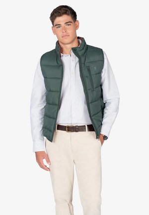 Chaleco verde acolchado con un cuello alto, cremallera frontal y bolsillos laterales. Lleva puesto una camiseta de rayas clara y se combina con pantalones beige.