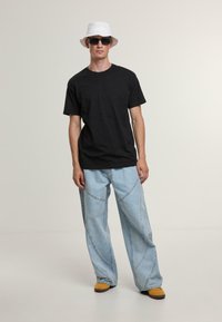 Schwarzes Baumwoll-T-Shirt, hellblaue weit geschnittene Jeans mit gebogenen Nähten, weißer Bucket Hat und schwarze Sonnenbrille. Gelbe Schuhe runden den Look ab.