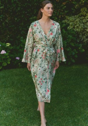 Woman walking barefoot on grass wearing a light green floral kimono robe tied at the waist in a garden setting.