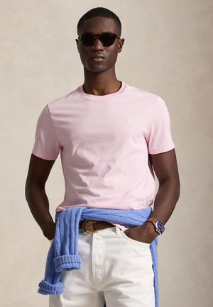 Mężczyzna w okularach przeciwsłonecznych, różowej koszulce, białych spodniach, niebieskim swetrze związanym wokół talii i niebieskim zegarku na lewym nadgarstku.