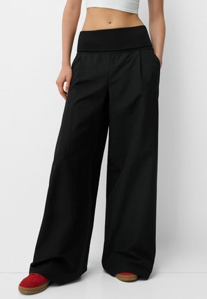 Frau trägt hochgeschnittene, weit geschnittene schwarze Hosen kombiniert mit roten Turnschuhen, steht vor einem schlichten weißen Hintergrund.