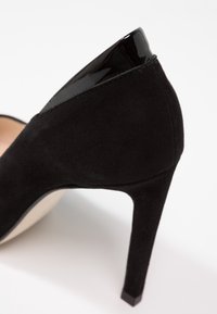 Chaussure à talons hauts noire avec extérieur en daim, accent en cuir verni brillant au talon et design effilé en stilettos.