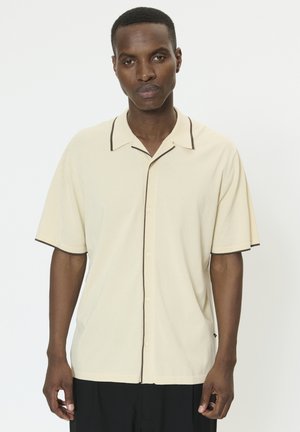 Beige skjorte med korte ærmer og brune kanter, knaplukning og blød tekstur. Har en klassisk krave og løs pasform.