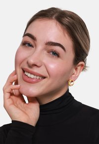 Zlatne naušnice u obliku obruča s uvijenim dizajnom. Model nosi crnu turtleneck majicu, smije se, ima svijetlu kožu i kosu srednje dužine.