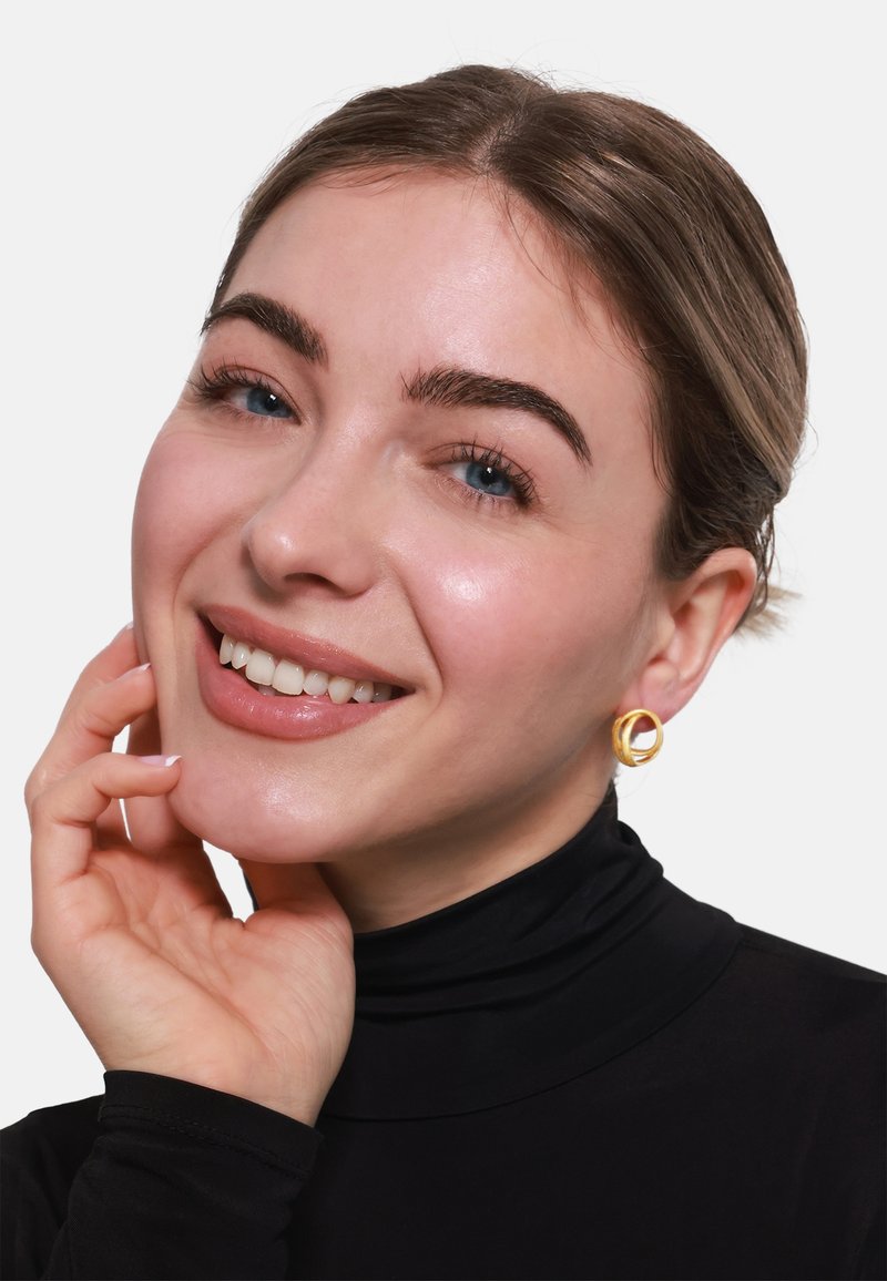 Zlatne naušnice u obliku obruča s uvijenim dizajnom. Model nosi crnu turtleneck majicu, smije se, ima svijetlu kožu i kosu srednje dužine.